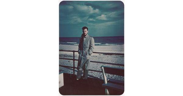 A color photograph of a White man in a long trench coat leaning against a railing with a beach in the background.