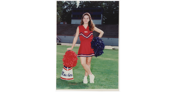 A color photograph of a young, smiling White woman in a red cheerleader uniform, holding pom-poms and leaning on a megaphone on a grassy sports field.