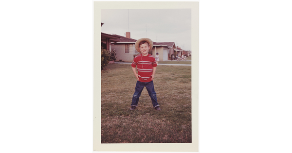 A color snapshot photograph of a young White child standing in a yard wearing a cowboy hat, red striped shirt, and blue jeans.