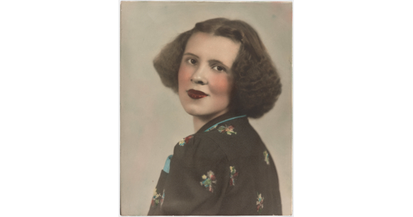 A color portrait photograph of a White woman with curly bobbed hair, wearing a floral top looking over her shoulder at the camera.