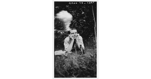 A black-and-white photograph of young White man in glasses, lying on his stomach in grass, his head propped in his hands; a beer bottle is in front of him, and a cloud of cigarette smoke hovers above his head.
