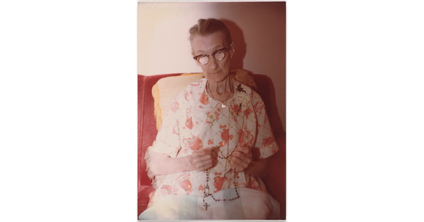 A color photograph of a seated elderly White woman, gray hair pulled back, wearing horn-rimmed glasses and a floral dress, a rosary in her hands.