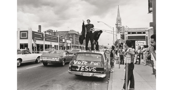 A black-and-white photograph of a downtown street scene of a man preaching from the top of a car with "Vote for Jesus" spray-painted on the back.