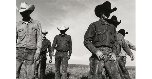 A black-and-white photograph of six cowboys walking in a field toward the viewer with their faces in shadow from their cowboy hats.