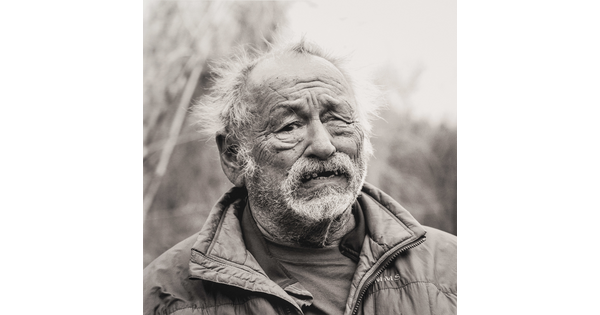 A black-and-white portrait photograph of a older light-skinned man, balding and with overgrown facial hair, wearing a quilted jacket standing outdoors.