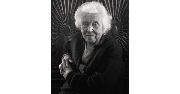 A black-and-white portrait photograph of an elderly White woman with fluffy white hair, posed in front of a carved wooden wall.
