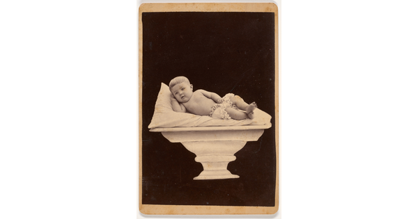 A black-and-white studio portrait photograph of a White baby in a diaper reclining on a pillow placed on a pedestal.