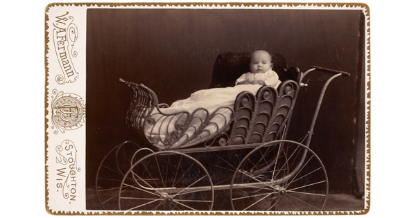 A black-and-white studio portrait photograph of a bald White baby posed on white pillows in an ornate baby buggy.