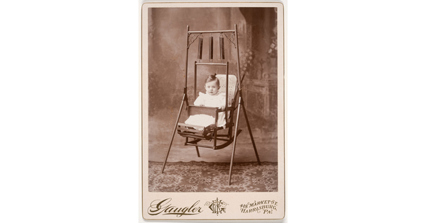 A black-and-white studio portrait photograph of a White toddler with dark hair sitting in a metal swing with three springs supporting the chair from the swing frame.