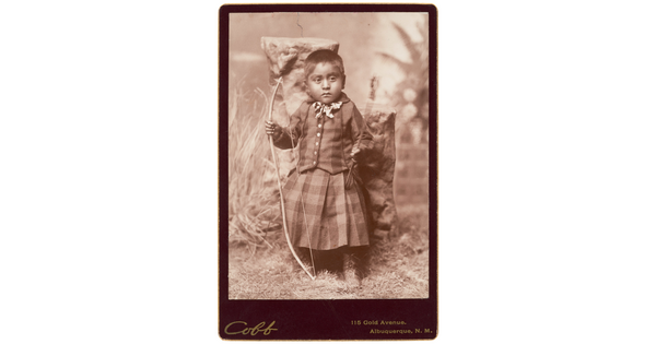 A sepia-toned studio portrait photograph of a young medium-skinned child with closely cropped dark hair wearing a tartan outfit and holding a bow and arrow.