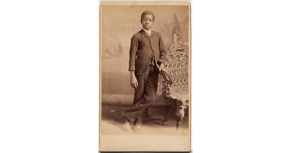A sepia-toned studio portrait photograph of a Black boy wearing a suit, leaning against a wrought iron bench.