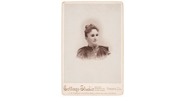 A black-and-white studio photograph of the head and shoulders of a White woman in Victorian-style dress wearing small, dark glasses.