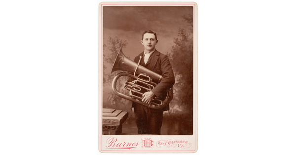 A black-and-white studio photograph of a White man in a suit holding a large trumpet-like brass instrument against his torso.