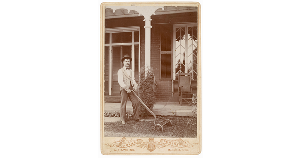 A black-and-white photograph of a White man with a mustache wearing bowler hat, bow tie, vest, and work gloves pushing a reel mower in front of a home.