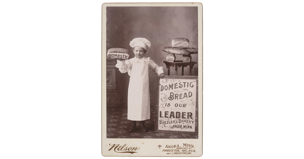 A black-and-white studio photograph of a White child dressed as a baker, holding up a loaf of bread that says "DOMESTIC" on its side, and standing next to a table of bread and a sign that reads, "Domestic bread is our leader, Brezler's Bakery, Anoka, Minn."
