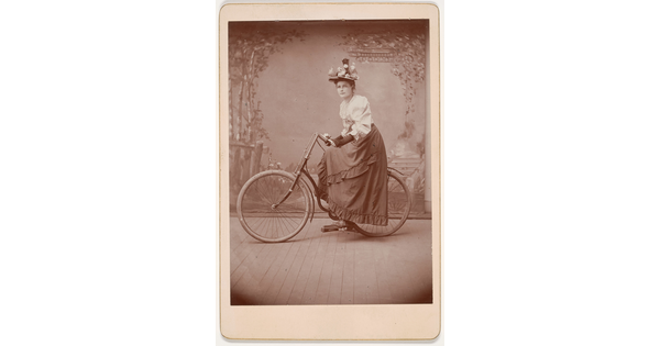 A black-and-white studio photograph of a White woman wearing a floral hat, white blouse, and long skirt posing on a bicycle in front of a scenic backdrop.