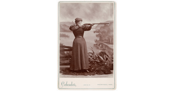 A black-and-white studio photograph of young White woman in a flat cap and plain, corseted gown aiming a rifle in front of a painted river scene.
