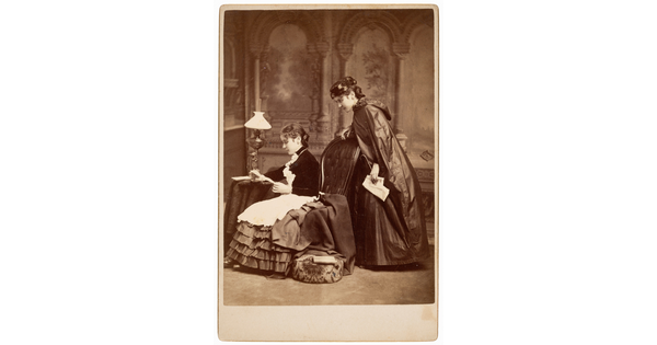 A black-and-white photograph of two identical White women in long dresses; one woman, seated, reads while the other stands behind and looks over her shoulder.