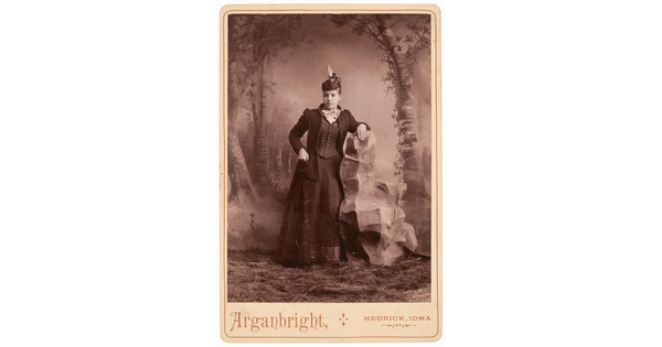 A sepia-toned studio photograph of a young White woman wearing a dark jacket, skirt and corseted vest, leaning on a fake rock in front of a painted forest landscape.