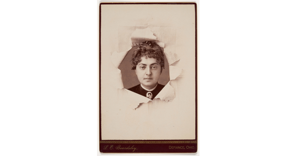 A black-and-white studio portrait photograph of a White woman behind paper that looks like it has been torn back to reveal her face.