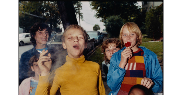 A color photograph of a group of smiling preteens: the boy in the front is smoking a cigarette and the one on the right is licking a lollipop.