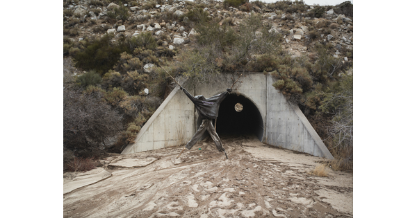 A color photograph of a human-like figure made from sticks and dressed in dark clothing in front of a drainage tunnel.