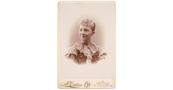 A sepia-toned studio portrait photograph of a White woman with curly, light colored hair in an updo, wearing a high-collared, frilly lace top.