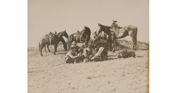 A sepia-toned photograph of a group of men sitting outside on the ground playing cards as their saddled horses stand behind them.