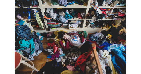 A color photograph of a light-skinned teenage girl lying on the floor of a messy room surrounded by unfolded clothing.