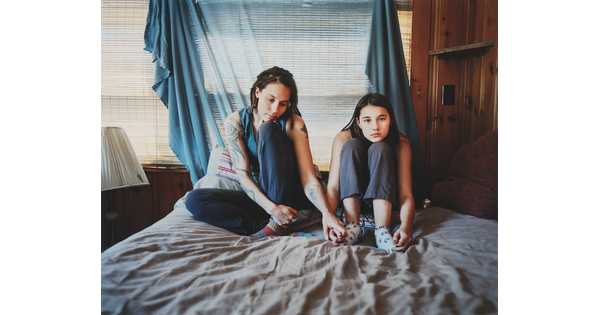 A color photograph of two light-skinned women holding hands and sitting on a bed in front of a fabric-covered window.