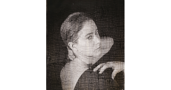 A black-and-white portrait photograph of a White woman, head turned to look over her shoulder, as viewed through what looks like a screen.