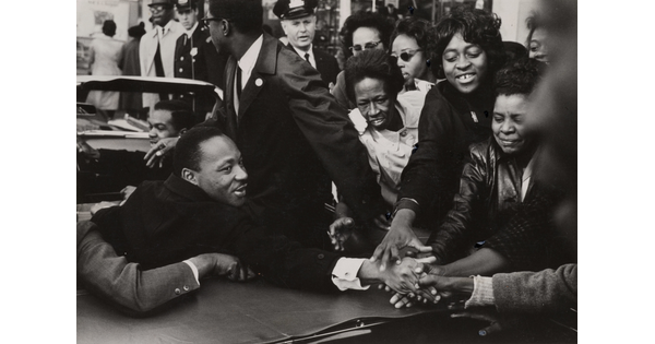 A black-and-white photograph of a Black man sitting in the back of a convertible and shaking hands with a crowd of Black people.