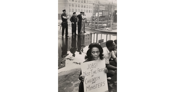 A black-and-white photograph of a Black woman holding a protest sign as White police officers stand in the background.
