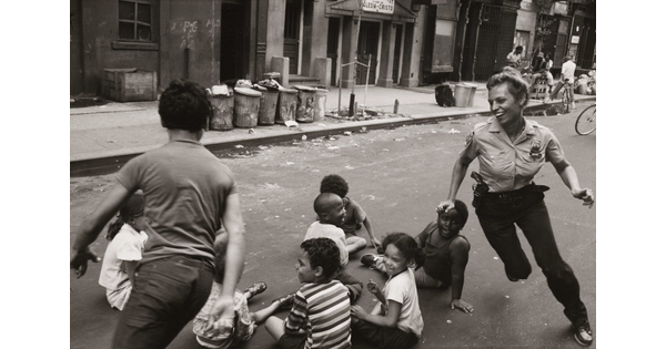 A black-and-white photograph of a smiling, White police woman playing a chase game with children of different races in a trash strewn street.