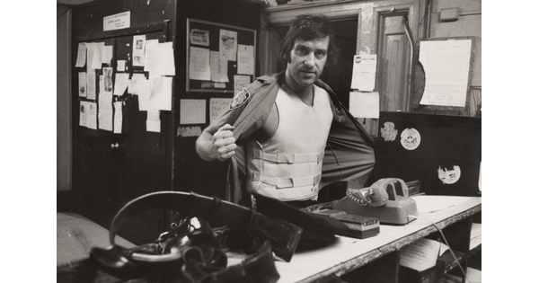 A black-and-white photograph of a White man standing behind a cluttered counter holding open his shirt to show a bullet-proof vest.