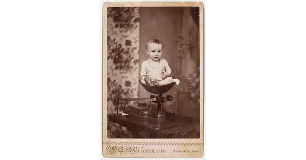 A sepia-toned studio photograph of a White baby wearing a necklace sitting in a scale.