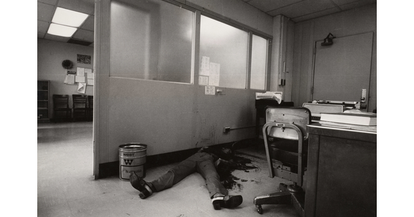 A black-and-white photograph of an office with a bloody body on the floor, feet toward the camera, the top part of the body obscured by dark stains on it, the floor, and the wall.