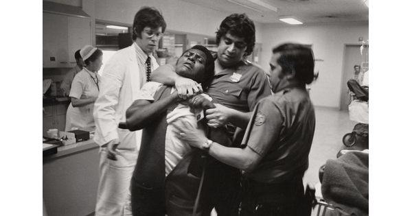 A black-and-white photograph of a Black man being restrained and choked by two police officers with medium to dark skin in a hospital setting.