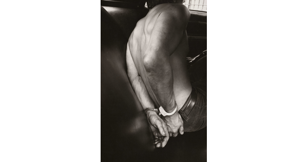 A black-and-white photograph of the bare back and handcuffed hands of a White man in the back seat of a police car.