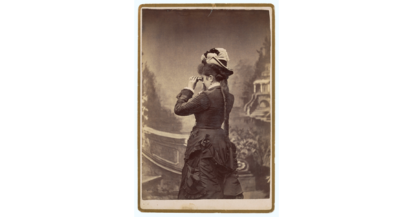 A sepia-toned studio photograph of a light-skinned woman, viewed from the back, wearing a dark corseted gown and peering through binoculars toward a painted landscape.