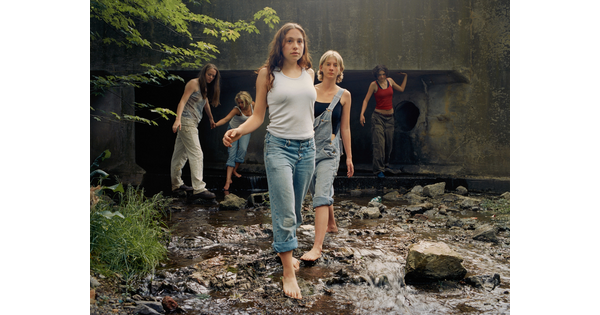 A color photograph of White teenagers, casually dressed, several barefoot, walking through a stream that emerges from a culvert behind them.