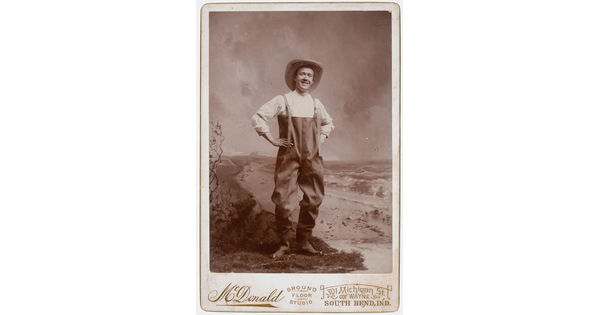A sepia-toned studio photograph of a smiling White man wearing waders and a large straw hat, posed with hands on hips.
