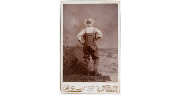 A sepia-toned studio photograph of a man, viewed from the back, wearing waders and a large straw hat, posed with hands on hips.