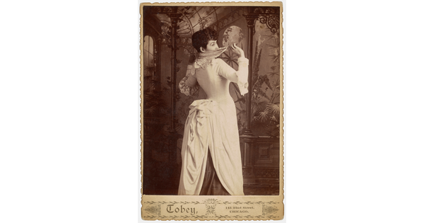 A sepia-toned studio photograph of a young White woman, viewed from the back, with a dramatically corseted dress and full bustle peeking playfully over her shoulder and hiding her face with a handkerchief.