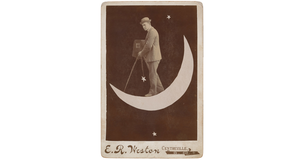 A black-and-white photograph of a White man wearing a bowler hat and suit holding an old-fashioned camera on a tripod while standing on a crescent moon.