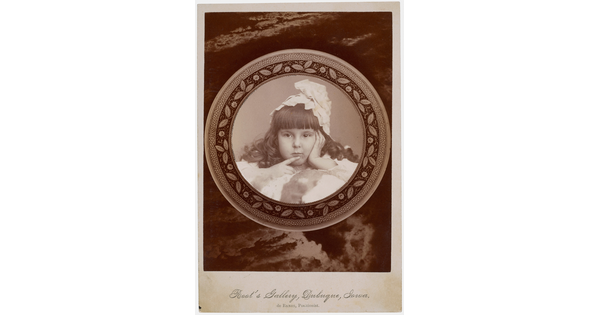 A black-and-white studio photograph of a young White girl with curled dark hair, bangs, and a massive bow on her head holding one finger to her mouth.