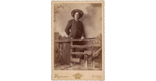 A sepia-toned photograph of a White man wearing a large sombrero-like hat standing behind a split-rail fence that is part of a studio set.
