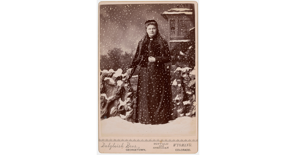 A black-and-white studio photograph of a White woman standing in a fake snowstorm against a backdrop of a winter scene.