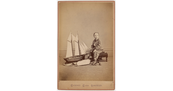 A sepia-toned studio photograph of a young White boy in a plaid suit seated on a bench next to a large toy sailboat.