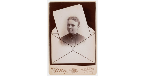 A sepia-toned studio photograph in which the head and shoulders of a White woman seems to be popping out of an envelope.